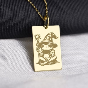 Può includere: Collana con pendente dorato. Il ciondolo rettangolare presenta un'illustrazione incisa di una rana mago, con cappello e veste, che tiene in mano un bastone. La catena della collana è dorata. La rana è vestita da mago.