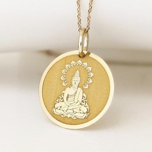 May include: A gold-colored circular pendant necklace featuring an engraved image of a seated figure in a meditative pose. The figure is surrounded by a halo of small petal-like shapes. The pendant hangs from a delicate gold-colored chain.
