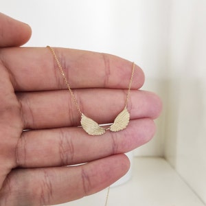 May include: A gold-toned necklace featuring a delicate chain and a pendant shaped like two symmetrical, textured angel wings. The wings are positioned horizontally, meeting in the center, and are suspended from a thin, polished gold-colored metal chain.