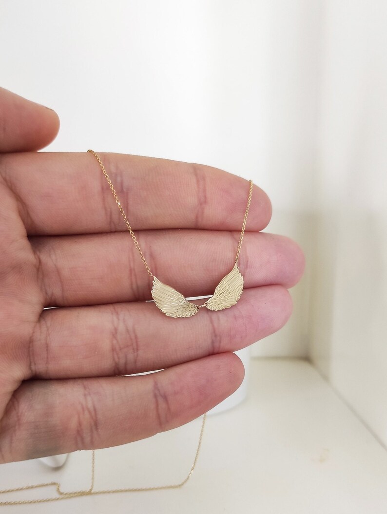 May include: A gold-toned necklace featuring a delicate chain and a pendant shaped like two symmetrical, textured angel wings. The wings are positioned horizontally, meeting in the center, and are suspended from a thin, polished gold-colored metal chain.