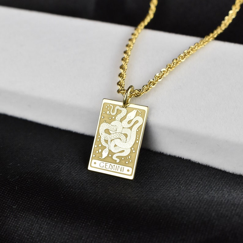 May include: Gold chain necklace with a rectangular pendant featuring a white etched design of two snakes intertwined. The pendant has the word "GEMINI" inscribed on it.