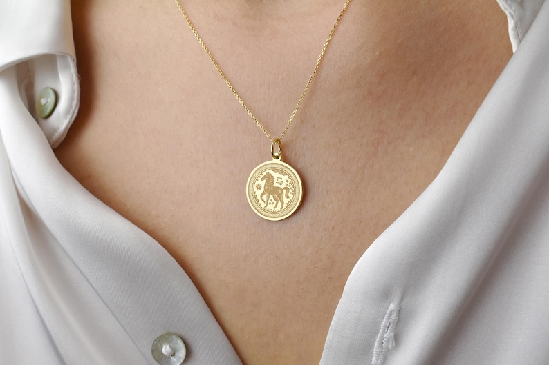 Peut inclure: Un collier en or avec un pendentif rond grav&eacute; d'un cheval et du caract&egrave;re chinois correspondant. Le m&eacute;daillon circulaire pr&eacute;sente une bordure d&eacute;corative et est suspendu &agrave; une fine cha&icirc;ne en or, reposant sur une chemise blanche &agrave; col.