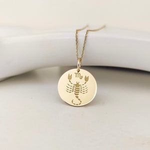 May include: A gold-colored circular pendant necklace featuring an engraved scorpion illustration and the Scorpio zodiac symbol. The pendant hangs from a delicate gold-colored chain, showcasing a polished metallic finish and a clean, flat design.