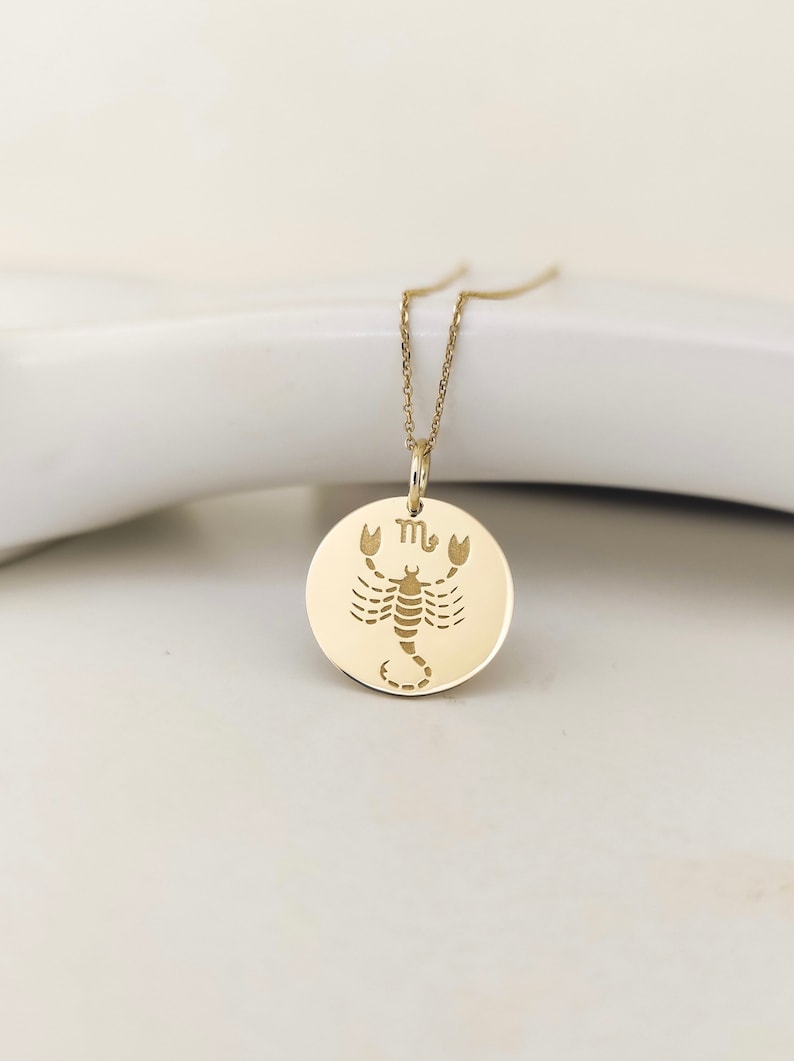 May include: A gold-colored circular pendant necklace featuring an engraved scorpion illustration and the Scorpio zodiac symbol. The pendant hangs from a delicate gold-colored chain, showcasing a polished metallic finish and a clean, flat design.