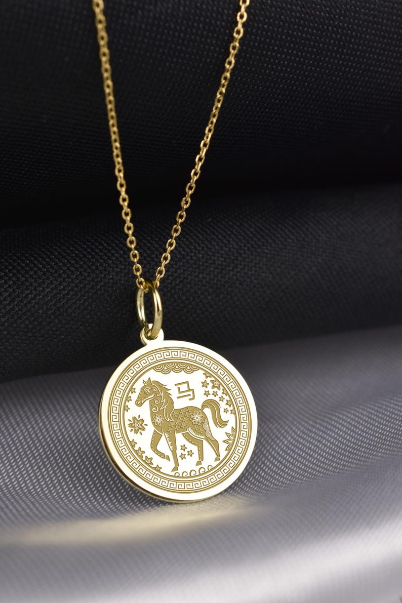 Peut inclure: Un collier pendentif circulaire dor&eacute; arborant un motif de cheval grav&eacute; au centre, accompagn&eacute; du caract&egrave;re chinois pour cheval, entour&eacute; d'une bordure g&eacute;om&eacute;trique d&eacute;corative et de petites &eacute;toiles, suspendu &agrave; une fine cha&icirc;ne dor&eacute;e.