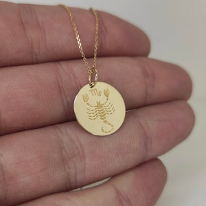 May include: A gold-colored necklace featuring a circular pendant with an engraved scorpion and the Scorpio zodiac symbol. The pendant hangs from a delicate gold-colored chain, showcasing a polished finish and detailed etching of the arachnid.