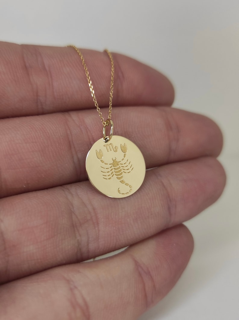 May include: A gold-colored necklace featuring a circular pendant with an engraved scorpion and the Scorpio zodiac symbol. The pendant hangs from a delicate gold-colored chain, showcasing a polished finish and detailed etching of the arachnid.