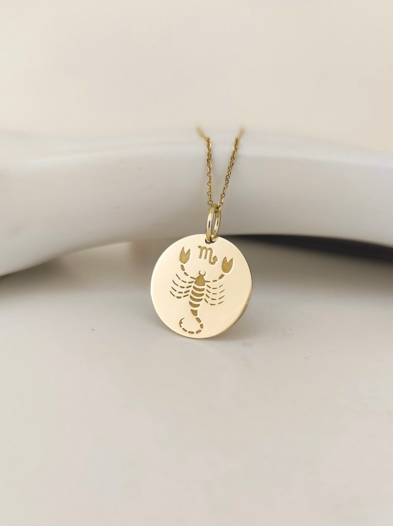 May include: A gold-toned circular pendant necklace featuring an engraved scorpion illustration and the Scorpio zodiac symbol. The pendant hangs from a delicate gold-toned chain, showcasing a polished metallic finish and a clean, flat circular design.