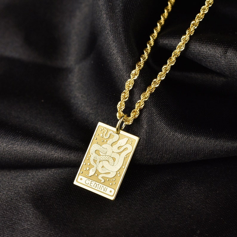 May include: Gold chain necklace with a gold pendant featuring a white outline of two snakes intertwined and the word "GEMINI" in white lettering.