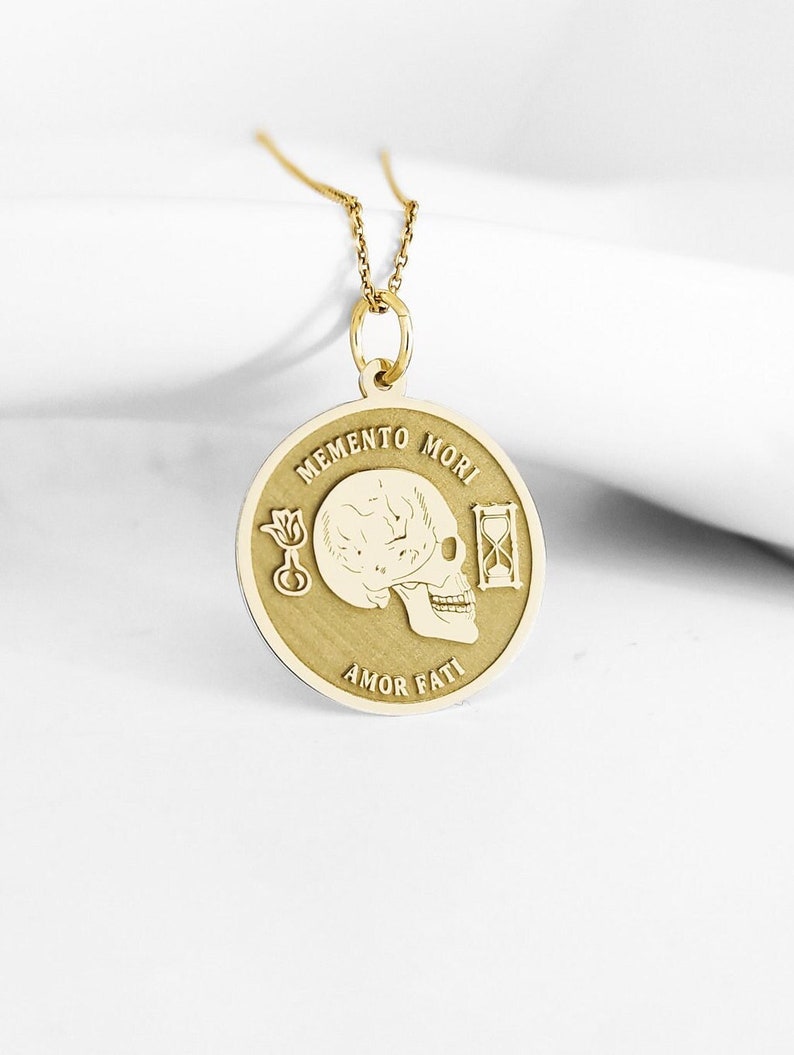 May include: A gold circular pendant necklace featuring an engraved human skull in the center, flanked by a small flower and an hourglass. The text MEMENTO MORI is inscribed above the skull, and AMOR FATI is inscribed below the skull design.