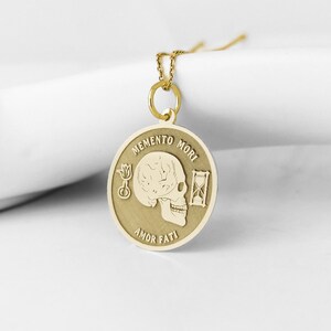 May include: A gold-toned circular pendant necklace featuring an engraved human skull in the center. The text MEMENTO MORI is inscribed above the skull, and AMOR FATI is inscribed below. A small flower and an hourglass are engraved on the sides.