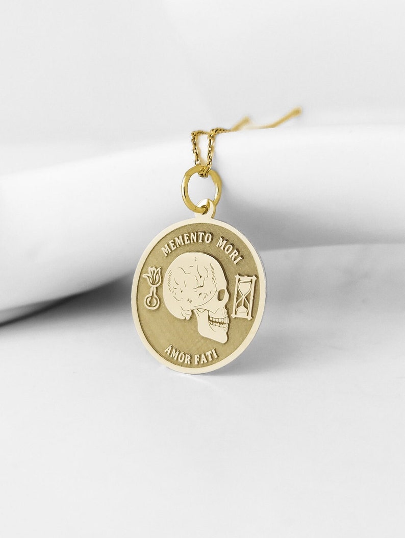May include: A gold-toned circular pendant necklace featuring an engraved human skull in the center. The text MEMENTO MORI is inscribed above the skull, and AMOR FATI is inscribed below. A small flower and an hourglass are engraved on the sides.