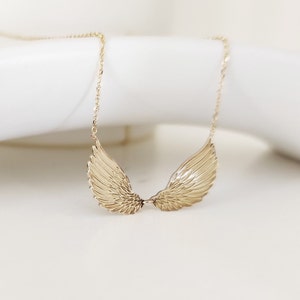 May include: A gold-toned necklace featuring a pendant shaped like two symmetrical angel wings. The wings are detailed with etched feather patterns and are connected at the center, suspended from a delicate, thin gold-toned metal chain.