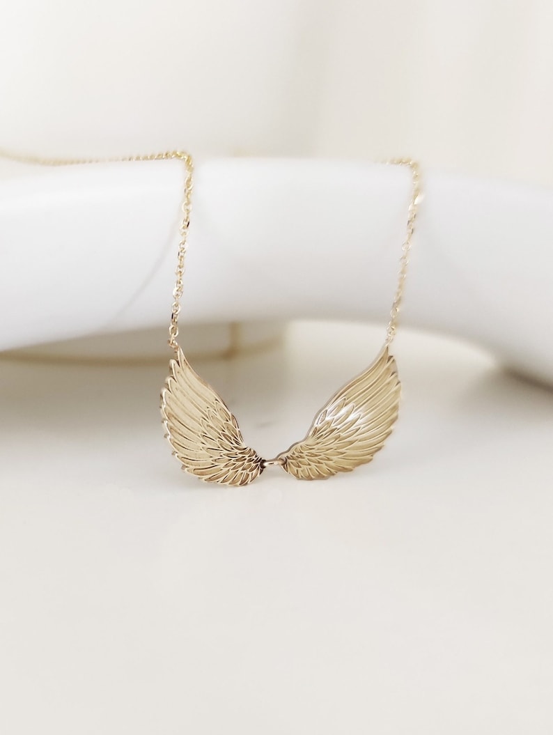 May include: A gold-toned necklace featuring a pendant shaped like two symmetrical angel wings. The wings are detailed with etched feather patterns and are connected at the center, suspended from a delicate, thin gold-toned metal chain.