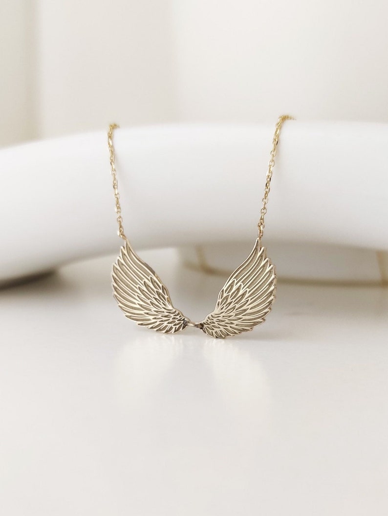 May include: A gold-toned necklace featuring a pendant shaped like two symmetrical, detailed angel wings. The wings are connected at the center and suspended from a delicate, thin gold-toned chain. The design showcases engraved feather textures.