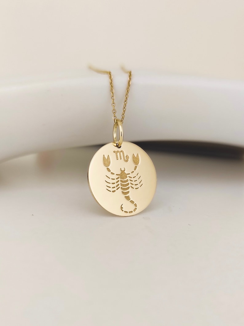 May include: A gold-toned circular pendant necklace featuring an engraved scorpion illustration and the Scorpio zodiac symbol at the top. The pendant hangs from a delicate matching gold-toned chain, showcasing a polished finish and clean lines.