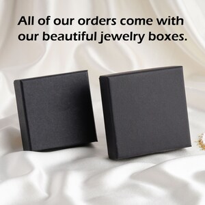 May include: Two square black jewelry boxes with a matte finish. The text "All of our orders come with our beautiful jewelry boxes." is printed in black above the boxes. The boxes are positioned upright, showing their front and side panels.