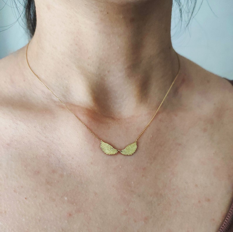 May include: A gold-toned necklace featuring a delicate chain and a central pendant shaped like two symmetrical, textured angel wings. The wings are positioned horizontally, meeting in the middle to form a single, cohesive winged design.