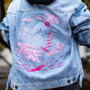 Peut inclure: Veste en jean bleu clair avec une capuche noire. Le dos de la veste présente un motif rose et blanc d'une montagne, de nuages et d'eau. La veste est portée sur un sweat à capuche noir.