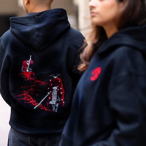 May include: Black hooded sweatshirts with red and white embroidered samurai designs. One sweatshirt features a large red emblem on the chest. The back of the other sweatshirt displays a detailed scene of a samurai warrior in a red robe with a sword.