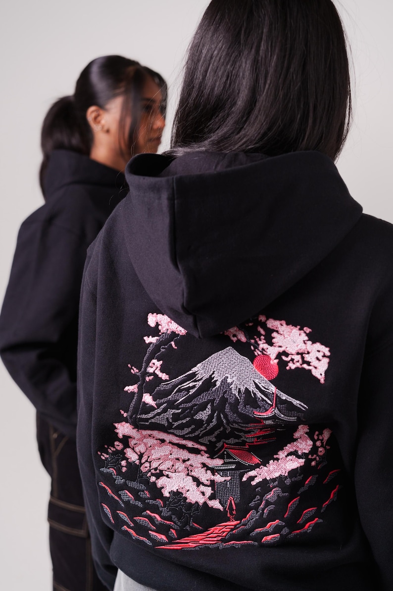 K&ouml;nnte beinhalten: Schwarzer Kapuzenpullover mit einem gestickten Design von Mount Fuji, Kirschbl&uuml;ten und einer roten Sonne. Das Design ist in Graut&ouml;nen, Rosa und Rot gehalten.