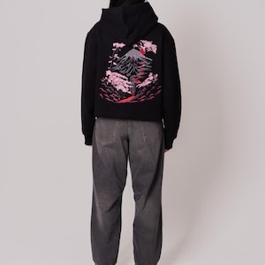 K&ouml;nnte beinhalten: Ein schwarzer Hoodie mit einem Aufdruck eines Berges und Kirschbl&uuml;ten auf der R&uuml;ckseite. Der Hoodie hat eine Kapuze mit Kordelzug und eine K&auml;ngurutasche.