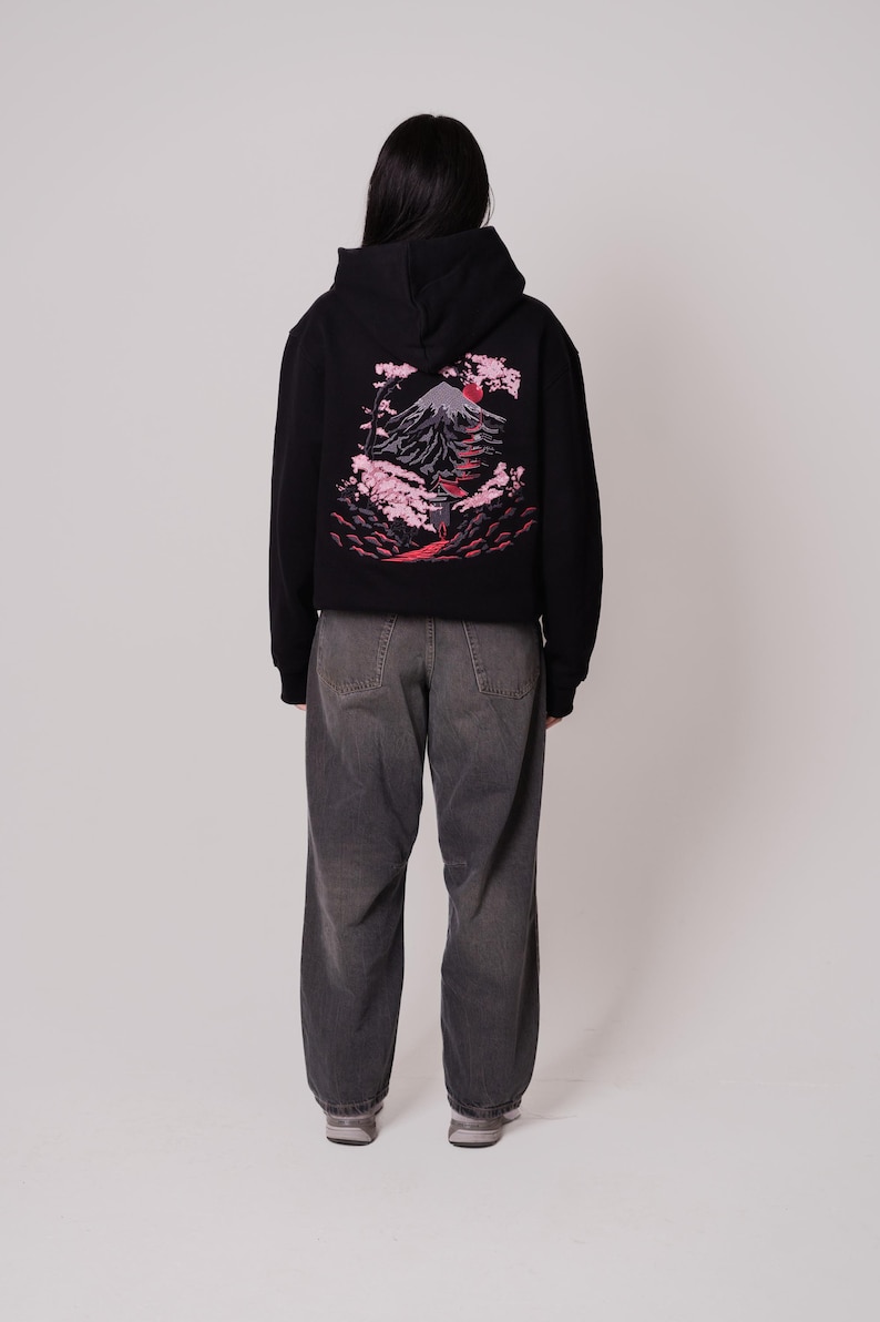 K&ouml;nnte beinhalten: Ein schwarzer Hoodie mit einem Aufdruck eines Berges und Kirschbl&uuml;ten auf der R&uuml;ckseite. Der Hoodie hat eine Kapuze mit Kordelzug und eine K&auml;ngurutasche.