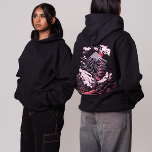 K&ouml;nnte beinhalten: Zwei Personen tragen schwarze Kapuzenpullover. Die Person rechts tr&auml;gt einen Kapuzenpullover mit einem gestickten Design in Rosa und Rot, das einen Berg und Kirschbl&uuml;ten auf der R&uuml;ckseite zeigt. Die Person links tr&auml;gt einen schlichten schwarzen Kapuzenpullover.