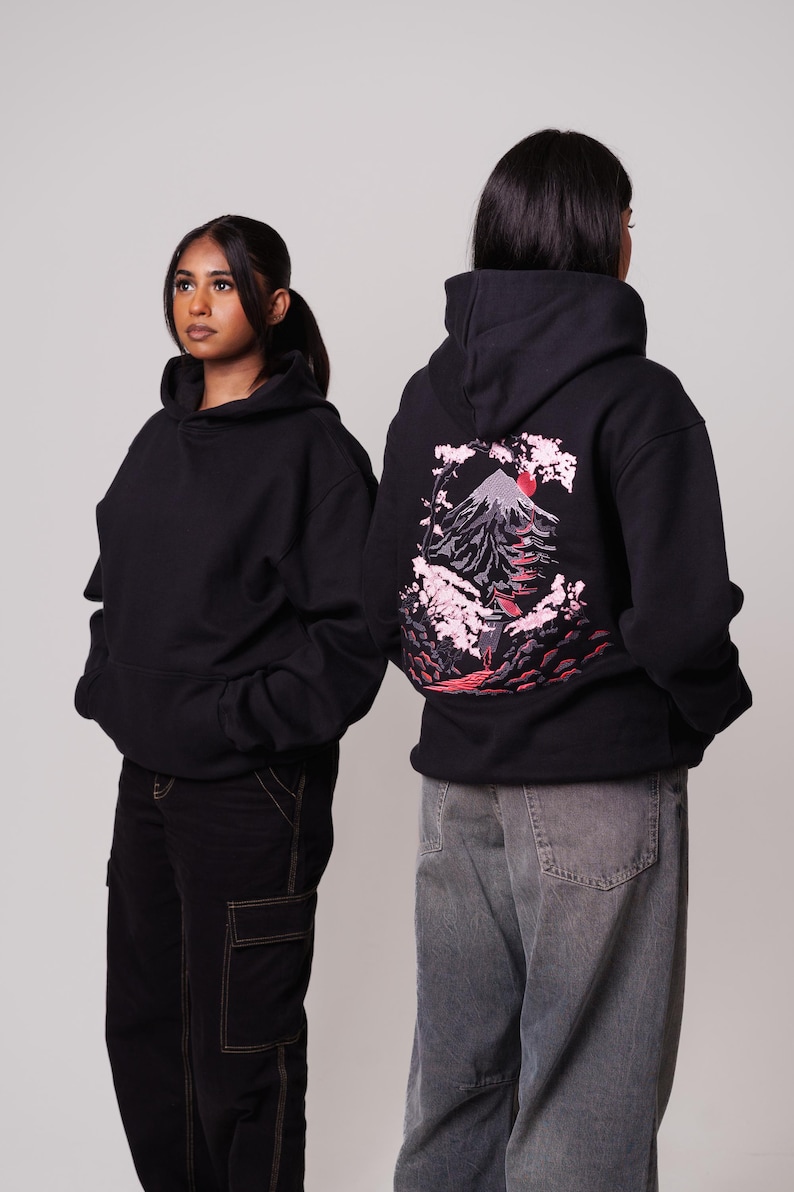 K&ouml;nnte beinhalten: Zwei Personen tragen schwarze Kapuzenpullover. Die Person rechts tr&auml;gt einen Kapuzenpullover mit einem gestickten Design in Rosa und Rot, das einen Berg und Kirschbl&uuml;ten auf der R&uuml;ckseite zeigt. Die Person links tr&auml;gt einen schlichten schwarzen Kapuzenpullover.