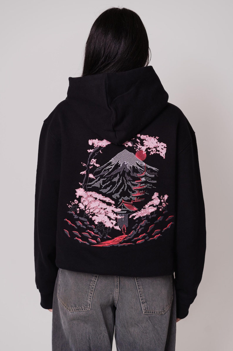 K&ouml;nnte beinhalten: Schwarzer Kapuzenpullover mit einem roten und rosafarbenen Stickmotiv von Mount Fuji, einem Pagodentempel und Kirschbl&uuml;ten.