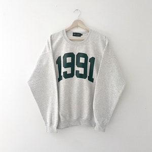 May include: A grey sweatshirt with a green 1991 printed on the front.