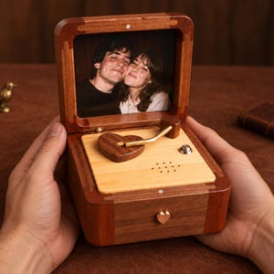 Puede incluir: Una caja de música de madera con una tapa abatible que muestra una fotografía de dos personas. La base interior es de madera clara con un componente en forma de corazón de madera oscura, un brazo dorado y un pequeño pomo en forma de corazón.