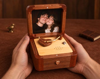 May include: A wooden music box featuring a hinged lid that displays a photograph of two people. The interior base is light-colored wood with a dark wood heart-shaped component, a gold-toned arm, and a small heart-shaped knob on the front.
