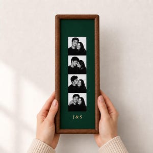 May include: A vertical rectangular wooden frame with a dark green mount containing a vertical strip of four black and white photo booth pictures of a couple. The text J & S is printed in gold lettering below the photos on the green mount.