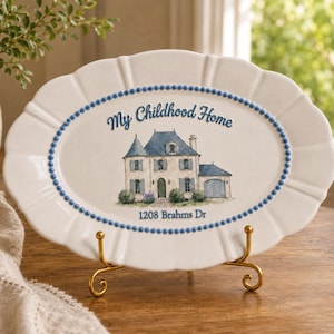 May include: An oval white ceramic platter with a scalloped edge and a blue beaded border. The centre features a watercolour illustration of a house with the text My Childhood Home and 1208 Brahms Dr. The platter rests on a gold metal stand.