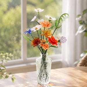 May include: A clear glass vase with an embossed pattern holds a bouquet of multi-colored glass flowers. The arrangement features orange, red, white, blue, and purple blossoms on thin green stems, creating a vibrant display of handcrafted glass art.