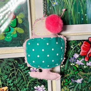 May include: A handmade fabric ice cream cone charm. The charm features a pink base, a teal polka-dot middle, and a white top with a pink pom-pom. It hangs from a silver chain. The background includes a painting of flowers and greenery.