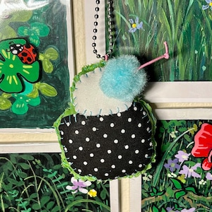 May include: Handmade fabric ornament shaped like a cupcake, featuring a black base with white polka dots, a white center, and a light blue pom-pom on top. The ornament has a green stitched edge and a silver chain for hanging.