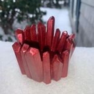 Puede incluir: Un cuenco rojo con forma de cristal. El cuenco está hecho de un material brillante y metálico y tiene una superficie rugosa y texturizada. El cuenco está sobre una superficie blanca con un fondo nevado.