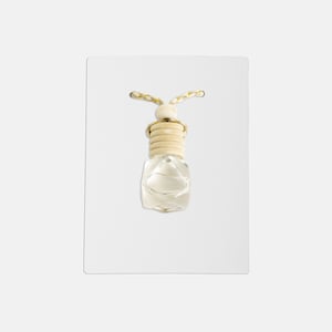 Puede incluir: Una botella de vidrio transparente con una tapa de madera y una cuerda dorada que cuelga de ella. La botella tiene forma de diamante y cuelga de una tarjeta blanca.