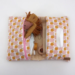 Peut inclure: Une pochette zippée blanche avec une fermeture éclair rose et un motif de petits cercles roses et jaunes. La pochette est ouverte et contient un animal en peluche, un jouet en bois et d'autres articles pour bébé. La pochette a un porte-mouchoirs sur le côté droit.