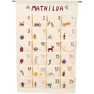 Può includere: Un calendario dell'avvento in tessuto color crema appeso a un'asta di legno, con il nome MATHILDA in alto. Presenta ventiquattro tasche individuali, numerate da uno a ventiquattro e decorate con illustrazioni colorate a tema natalizio.
