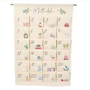 Pode incluir: Um calendário do advento de tecido na cor creme pendurado numa haste de madeira, com o nome Mathilda no topo. Possui vinte e quatro bolsos individuais, cada um numerado e decorado com pequenas ilustrações coloridas com temas festivos, como árvores, presentes e animais.