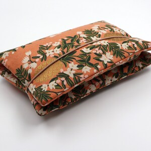 May include: A peach-colored fabric pouch with a floral print featuring white flowers and green leaves. The pouch has a brown strap and is folded closed.