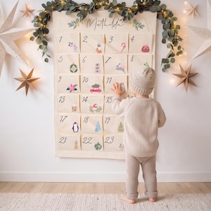 Può includere: Un calendario dell'avvento in tessuto color crema appeso a una parete, con il nome Mathilda scritto in corsivo nero nella parte superiore. Il calendario presenta ventitré tasche numerate, ognuna decorata con piccole illustrazioni natalizie.
