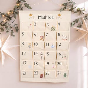 Pode incluir: Um calendário do advento de tecido na cor creme com o nome Mathilda no topo, vinte e quatro bolsos numerados e pequenas ilustrações em aquarela, incluindo um presente, um cavalo de balanço, um quebra-nozes e uma árvore de Natal.