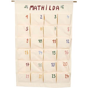 Pode incluir: Um calendário do advento de tecido creme com 24 bolsos numerados. O nome "MATHILDA" é bordado no topo, com folhas verdes decorativas. Os números são em várias cores, incluindo vermelho, azul e verde.