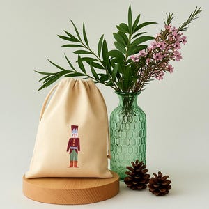 Op de afbeelding: Een beige trekkoordtas met een geborduurd speelgoedsoldaatontwerp. De tas staat op een houten standaard, naast een groene glazen vaas met bloemen en groen. Twee dennenappels staan aan de voet van de vaas.