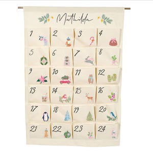 Può includere: Un calendario dell'avvento in tessuto color crema appeso a un'asta di legno, con il nome Mathilda in alto. Presenta ventiquattro tasche individuali, ciascuna numerata e decorata con piccole illustrazioni colorate a tema natalizio.