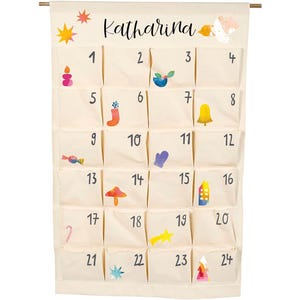 Könnte beinhalten: Ein cremefarbener Adventskalender aus Stoff, der an einer Holzstange hängt und oben den Namen Katharina trägt. Er verfügt über vierundzwanzig nummerierte Taschen, die jeweils mit kleinen, bunten, abstrakten Illustrationen weihnachtlicher Motive verziert sind.
